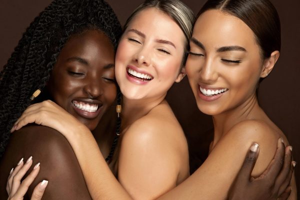 Three diverse women embracing with eyes closed and smiling, celebrating friendship and unity.